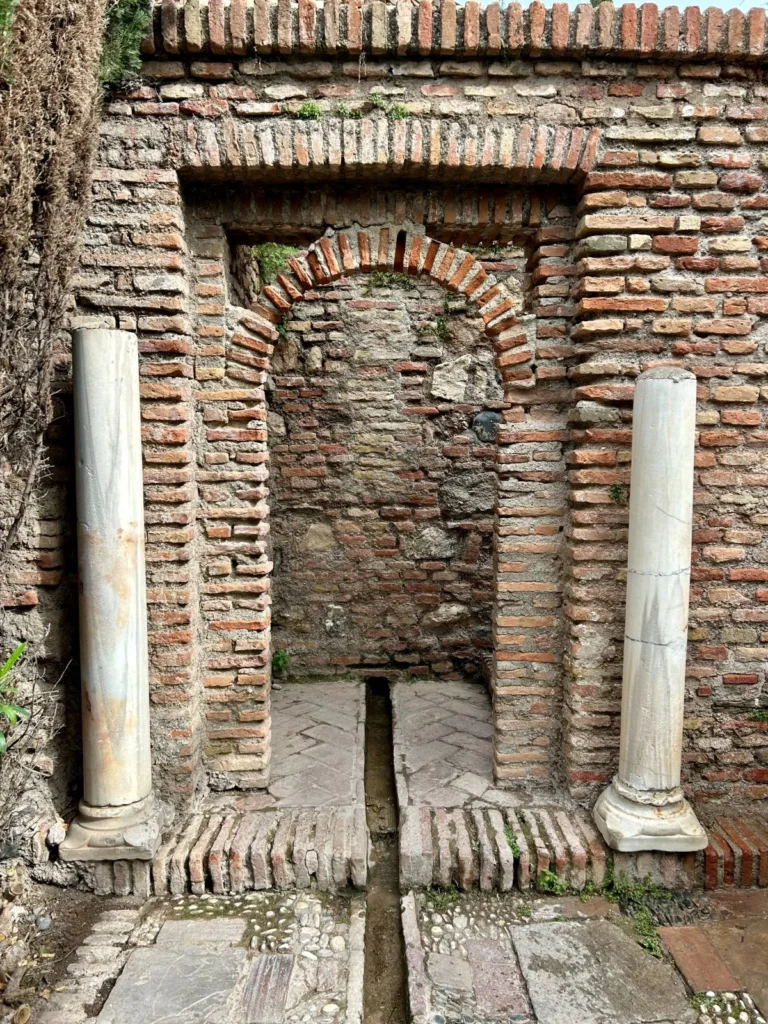 Ruined wall fountain niche at the Alcazaba Málaga, framed by two reused Roman marble columns and a brick horseshoe arch, with a water channel running along the stone floor