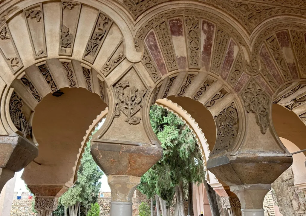 Underside of ornately carved stucco horseshoe arches at the Alcazaba palace in Málaga, showing intricate vegetal and geometric relief work with traces of original red paint and cypress trees visible through the openings