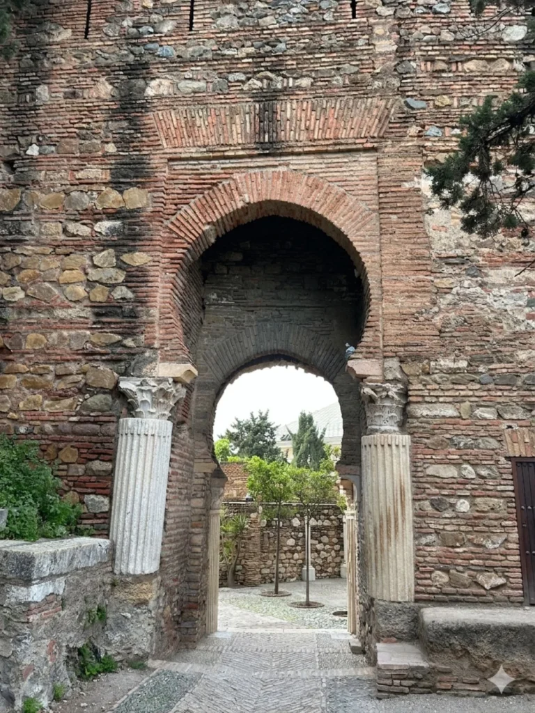 Monumental entrance gateway of the Alcazaba in Málaga at the beginning of the free tour, showing a tall horseshoe brick arch flanked by two reused Roman fluted marble columns with Corinthian capitals, opening onto a cobblestone passage leading into the fortress interior