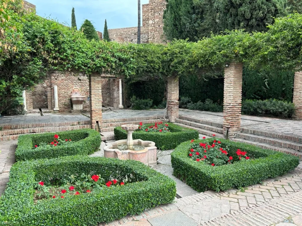 Formal Moorish garden courtyard at the Alcazaba in Málaga, featuring a carved stone fountain surrounded by neatly trimmed box hedges and red geraniums, with ancient brick columns and cypress trees in the background on alcazaba free tour