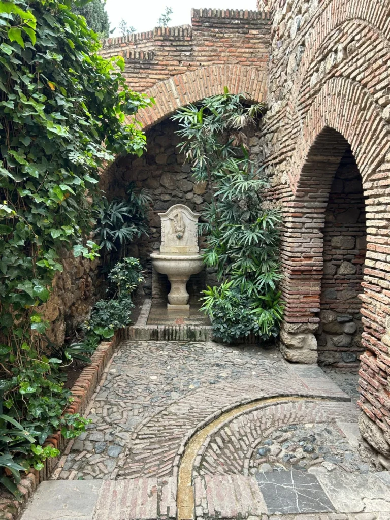 Secluded garden passage inside the Alcazaba, Málaga, with a carved stone wall fountain set against lush tropical planting, framed by pointed brick arches and ivy-covered walls with a mosaic cobblestone floor
