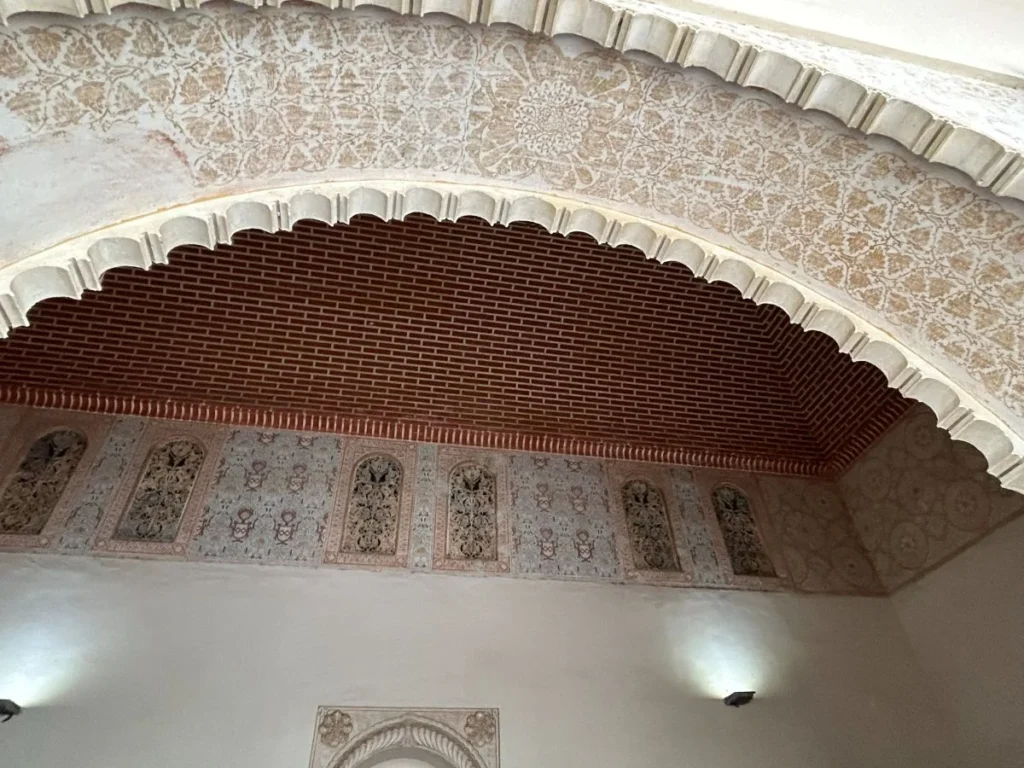 Close-up of a scalloped Moorish arch inside the Alcazaba, Málaga, showing intricate gold-and-white stucco arabesques on the ceiling, brick intrados, and painted decorative panel niches below