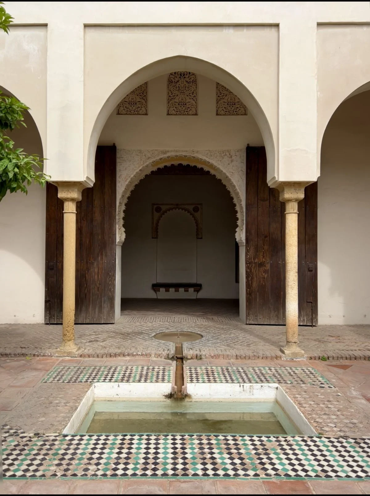 Symmetrical view of the Nasrid palace inner courtyard at the Alcazaba in Málaga, featuring a rectangular pool edged in green, black and white zellige diamond-pattern tiles, a central fountain jet, triple arched portico on stone columns, carved wooden doors, and a layered Moorish horseshoe arch leading to an inner chamber