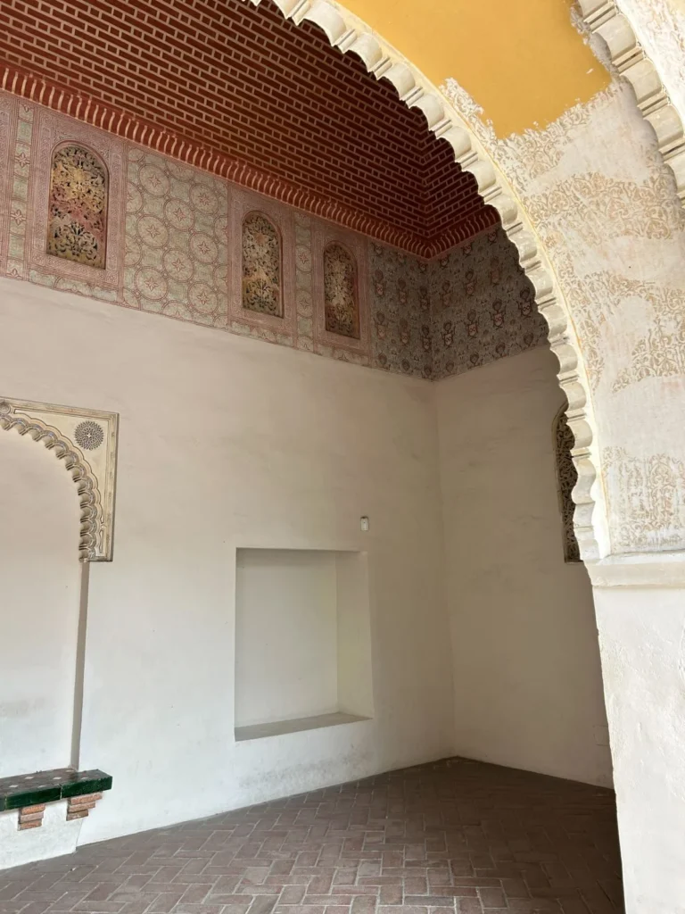 Interior corner of a Nasrid palace chamber at the Alcazaba in Málaga, showing a colourful painted stucco frieze with floral and geometric panels, a scalloped brick cornice, a large Moorish arch with arabesques, and a recessed wall niche