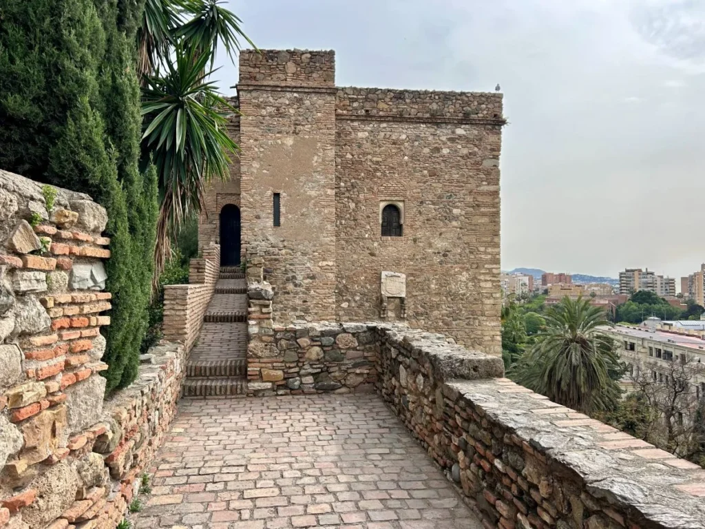 Exterior of a large stone and brick defensive tower at the Alcazaba in Málaga, showing narrow arrow-slit windows, a small Moorish arched window, and a carved stone heraldic coat of arms plaque on the wall, with palm trees and city views beyond