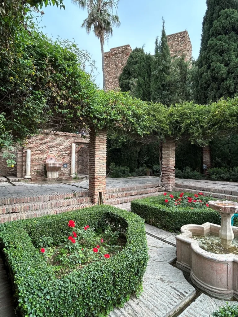 The Patio de Armas at the Alcazaba in Málaga, showing a stone fountain surrounded by clipped box hedges and red geraniums, brick pergola columns draped with climbing vines, a wall fountain niche with Roman columns, and a Moorish tower rising above the cypress trees