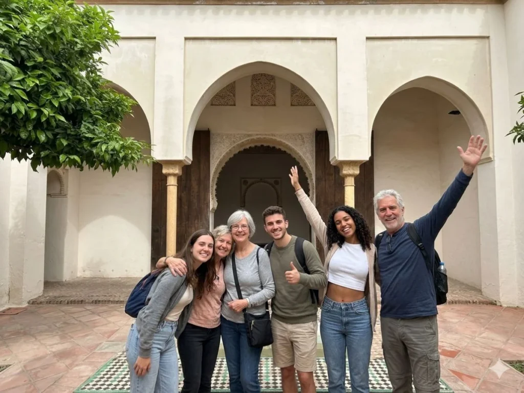Six happy tourists of mixed ages smiling and waving inside the Nasrid palace courtyard of the Alcazaba in Málaga, standing in front of the triple horseshoe arch portico with carved wooden doors and zellige tile pool during a guided private tour