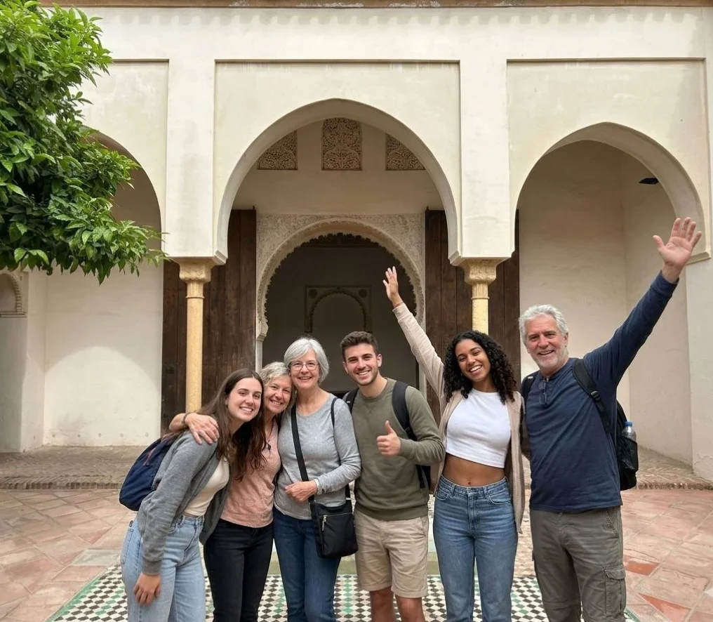 Six happy tourists of mixed ages smiling and waving inside the Nasrid palace courtyard of the Alcazaba in Málaga, standing in front of the triple horseshoe arch portico with carved wooden doors and zellige tile pool during a guided private tour