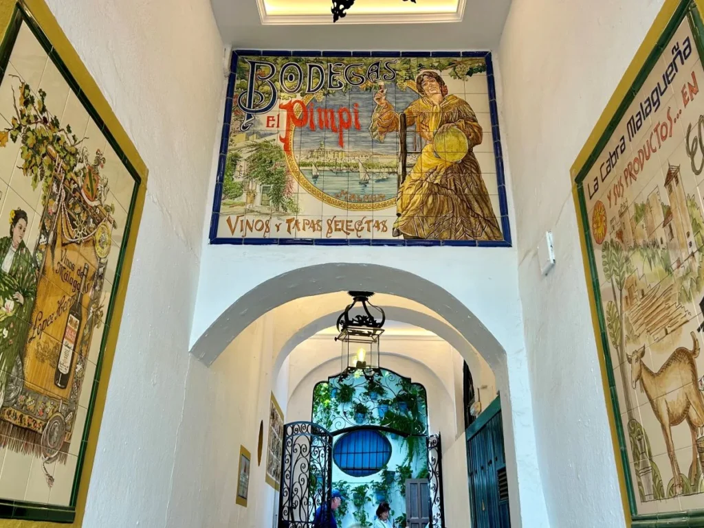 Entrance hallway of Bodega El Pimpi in Málaga, looking through a white arched corridor toward the plant-filled patio, with the iconic hand-painted ceramic tile mural reading "Bodega El Pimpi — Vinos y Tapas Selectas" mounted above the arch and decorative wine murals on either side