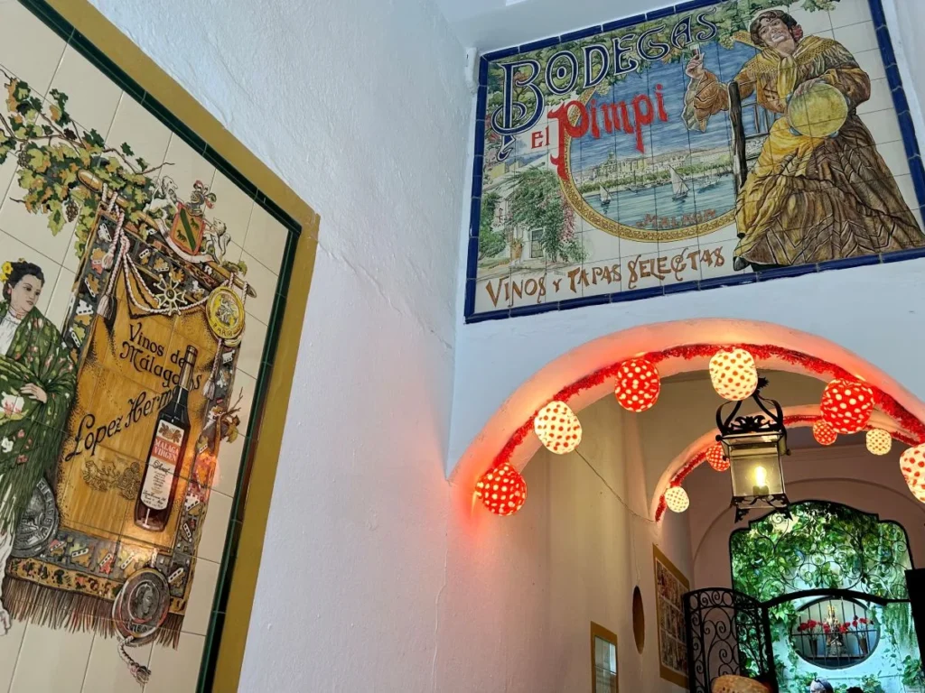Evening view of the Bodega El Pimpi hallway in Málaga, with the iconic ceramic tile mural above a Moorish arch decorated with red and white polka dot Feria lanterns, an ornate López Hermanos wine mural to the left, and the lit garden patio visible through the wrought iron gate