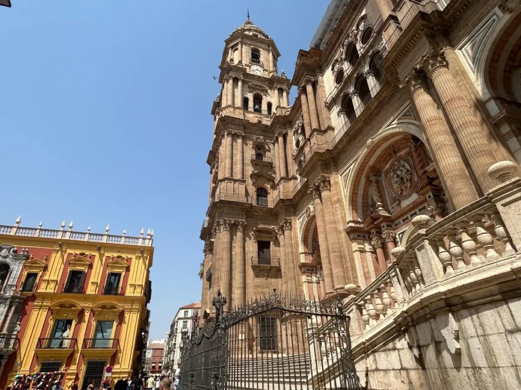 Looking up at the soaring baroque bell tower and richly carved stone facade of Málaga Cathedral from the plaza below, with the ornate iron entrance gates and balustrade staircase in the foreground and the colourful yellow Bishop's Palace visible to the left