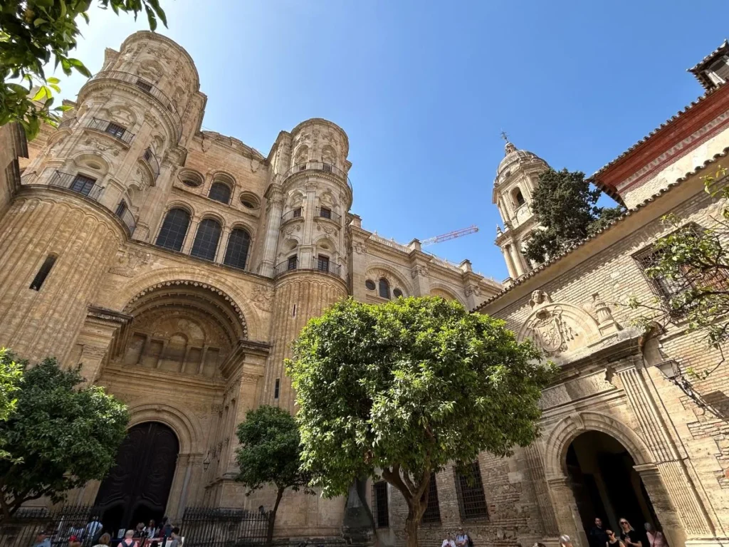 Looking up at the imposing baroque facade of Málaga Cathedral from the orange tree courtyard, showing the ornately carved main doorway arch, the massive ribbed drum towers rising above, the completed bell tower to the right, and visitors seated at the base on a sunny day