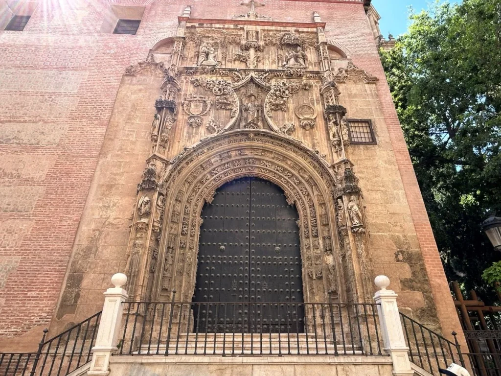 Close-up of the Puerta del Sagrario on the side of Málaga Cathedral, featuring an extraordinarily detailed Plateresque carved stone doorway with saints, heraldic shields, and religious figures surrounding tall iron-studded black wooden doors set into a brick wal