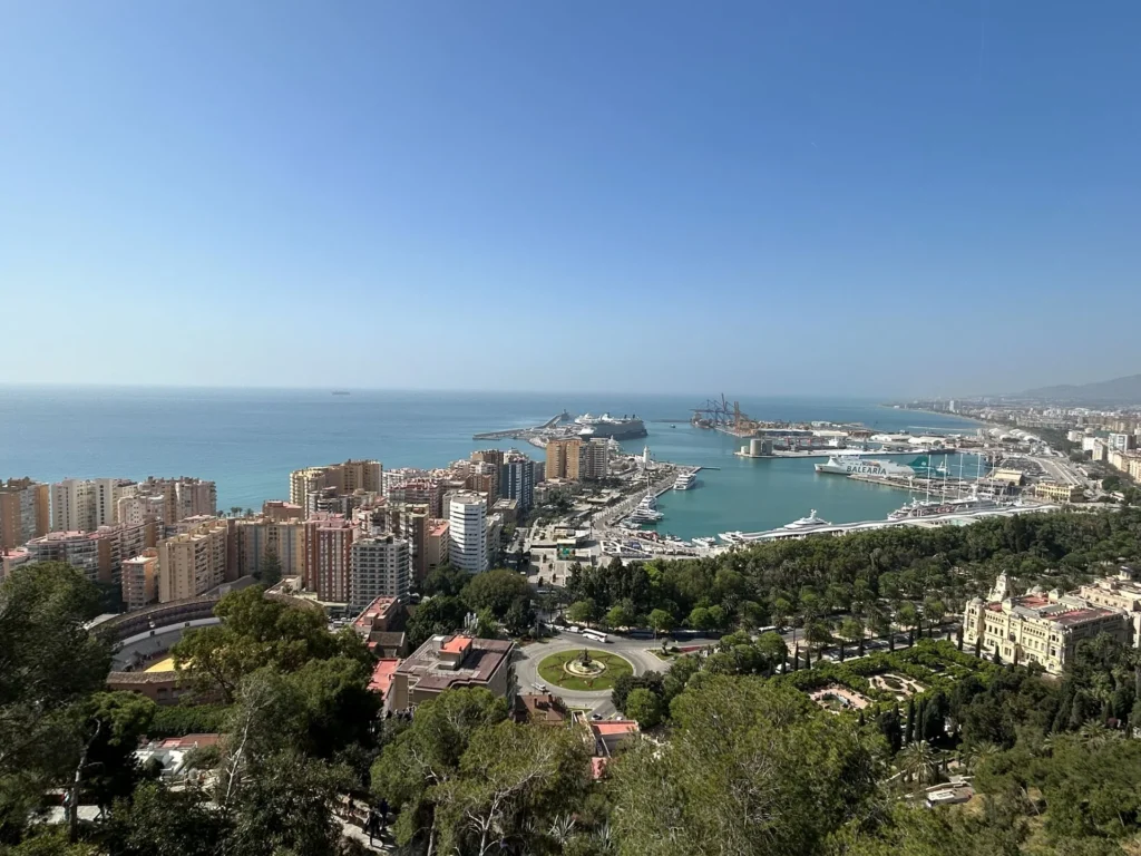 Sweeping panoramic view from the Alcazaba hilltop in Málaga during a free tour, looking over the city rooftops, the Parque de Málaga, the city hall building, and the curved port with cruise ships docked along the Mediterranean coastline
