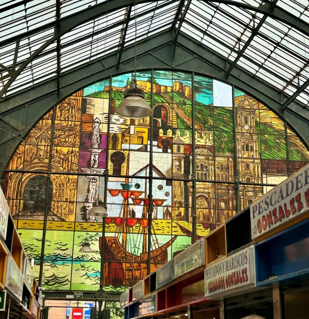Interior view of the Mercado Central de Atarazanas in Málaga, looking up at the vast iron and glass barrel-vaulted roof above a giant stained glass window depicting the Alcazaba, Málaga Cathedral, tall ships, and the city's history, with market stall signs visible below