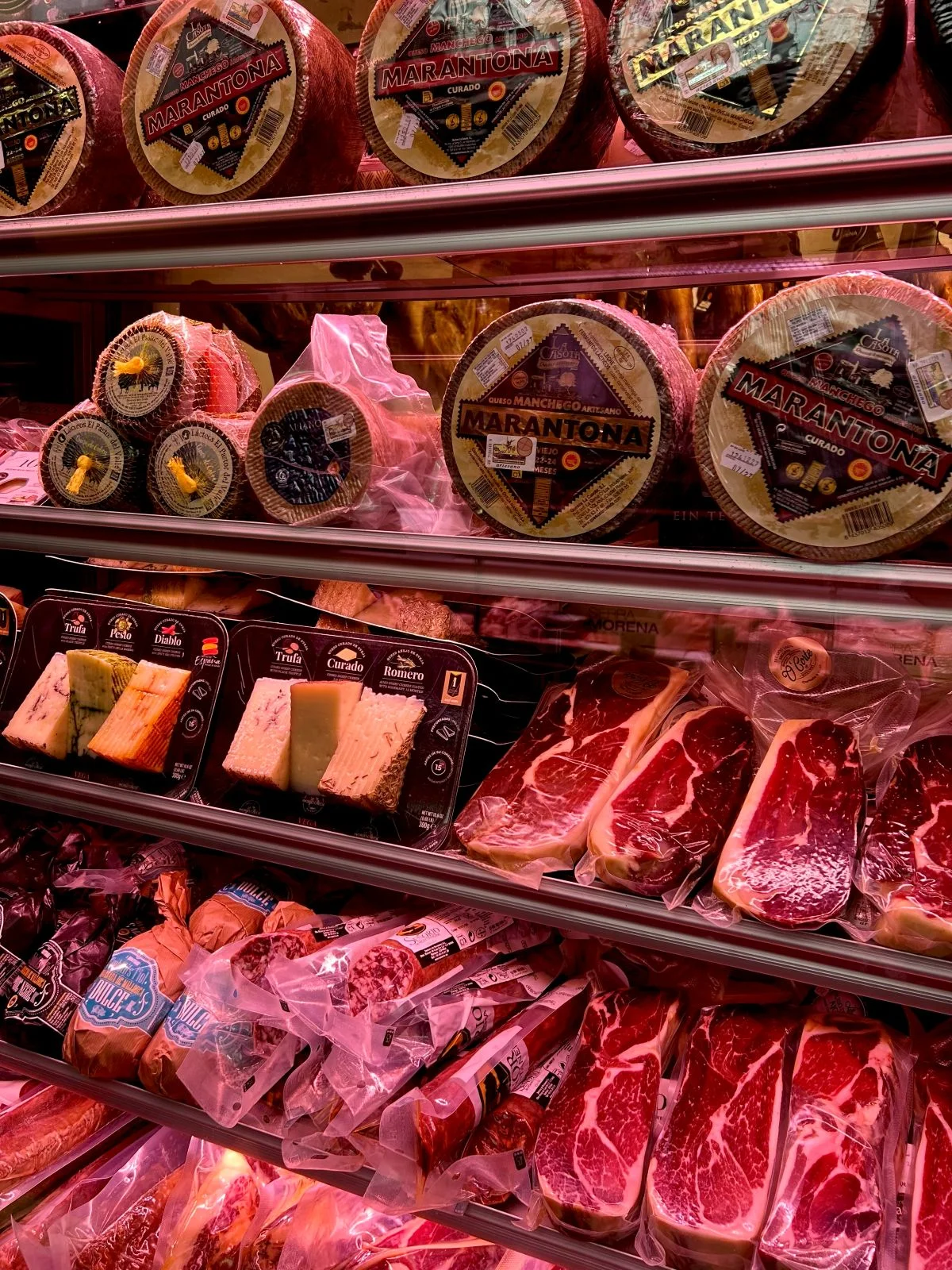 Refrigerated market stall display at the Mercado de Atarazanas in Málaga, packed with whole wheels of Marantona Manchego cheese, Ibérico ham cuts, and assorted Spanish cured meats under pink display lighting