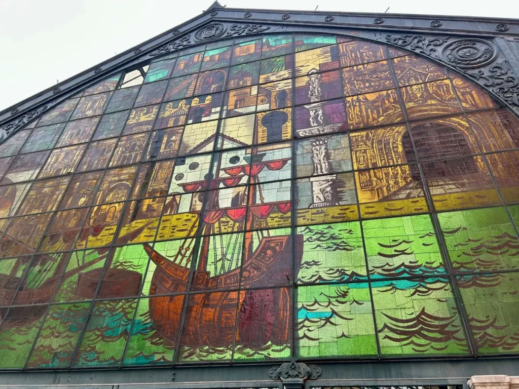 Close-up of the full stained glass gable facade of the Mercado de Atarazanas in Málaga, showing a large tall ship at sea surrounded by depictions of Málaga's landmarks including the Alcazaba and cathedral, set within an ornate iron-framed market building exterior