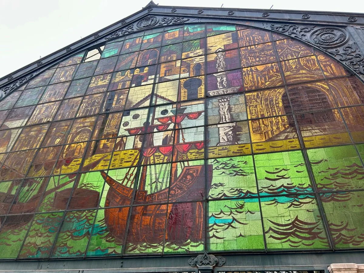 Close-up of the full stained glass gable facade of the Mercado de Atarazanas in Málaga, showing a large tall ship at sea surrounded by depictions of Málaga's landmarks including the Alcazaba and cathedral, set within an ornate iron-framed market building exterior