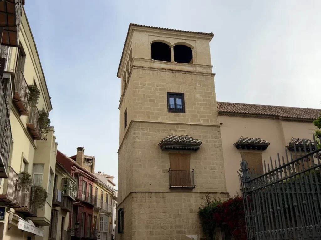 A plain ashlar stone church tower rising above a narrow street in Málaga's old town, with open belfry arches at the top, traditional balconied apartment buildings alongside, terracotta roof tiles, and iron railings with climbing red flowers at the base