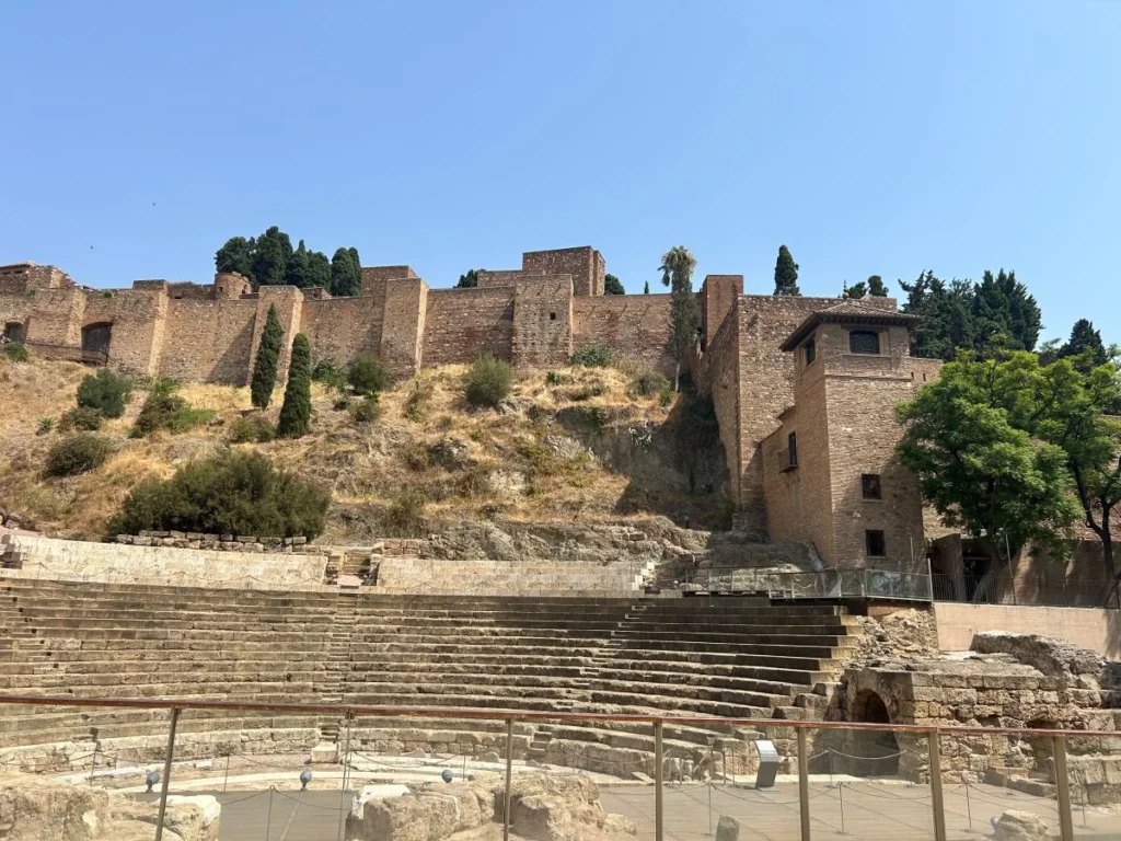 Ancient Roman theatre ruins in Málaga showing stone seating tiers and excavated foundations, with the Moorish Alcazaba fortress and its brick tower rising directly above on the hillside during alcazaba and roman theatre free tour