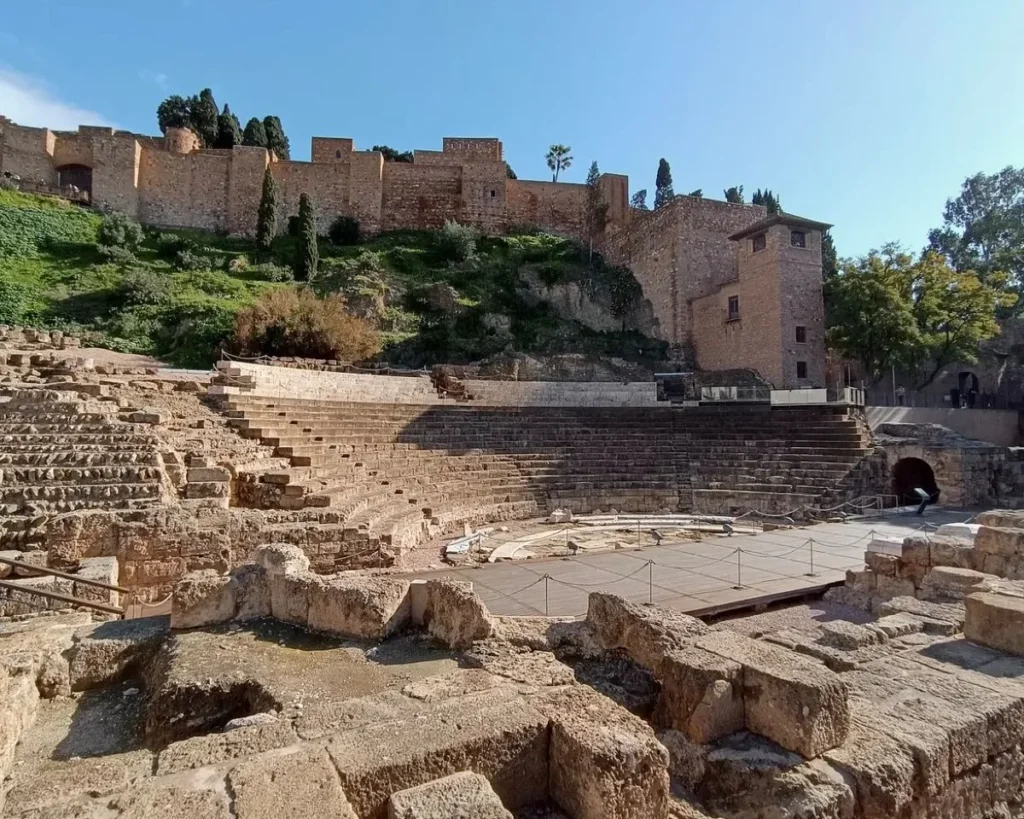 Ground-level view of the Roman theatre in Málaga showing the semicircular stone seating tiers, excavated orchestra pit, and reconstructed stage platform, with the full Alcazaba Moorish fortress walls and towers dominating the green hillside directly above