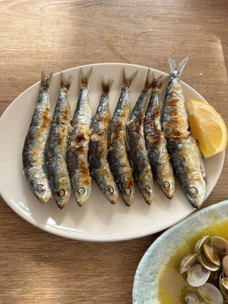 A plate of freshly grilled sardines espetos with sea salt and lemon wedge alongside a bowl of clams in broth, classic Málaga seafood tapas served at a local restaurant