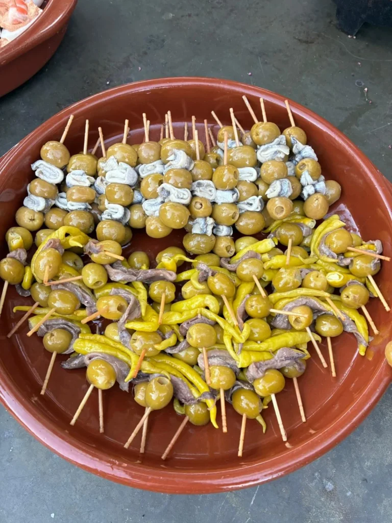 A terracotta dish filled with two styles of banderilla skewers — green olives stuffed with blue cheese, and green olives with anchovies and pickled yellow guindilla peppers — a classic Spanish tapas bar snack in Málaga