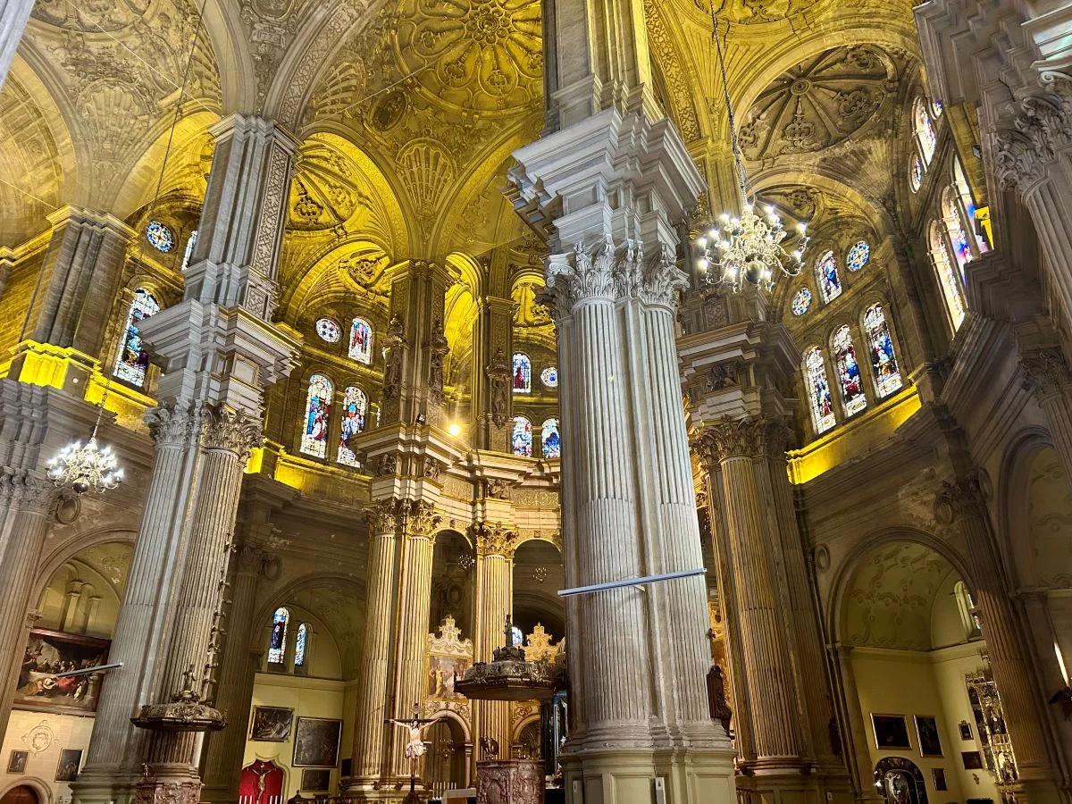 Wide view of Málaga Cathedral's interior with tall Corinthian columns, chandeliers and the gilded altar in the background